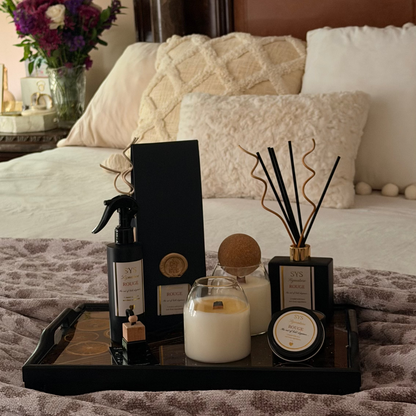 A styled bedroom scene with a bed in the background and a black tray in the foreground holding a candle, a reed diffuser, a small bottle, and a rectangular box. The tray rests on a leopard-print blanket.