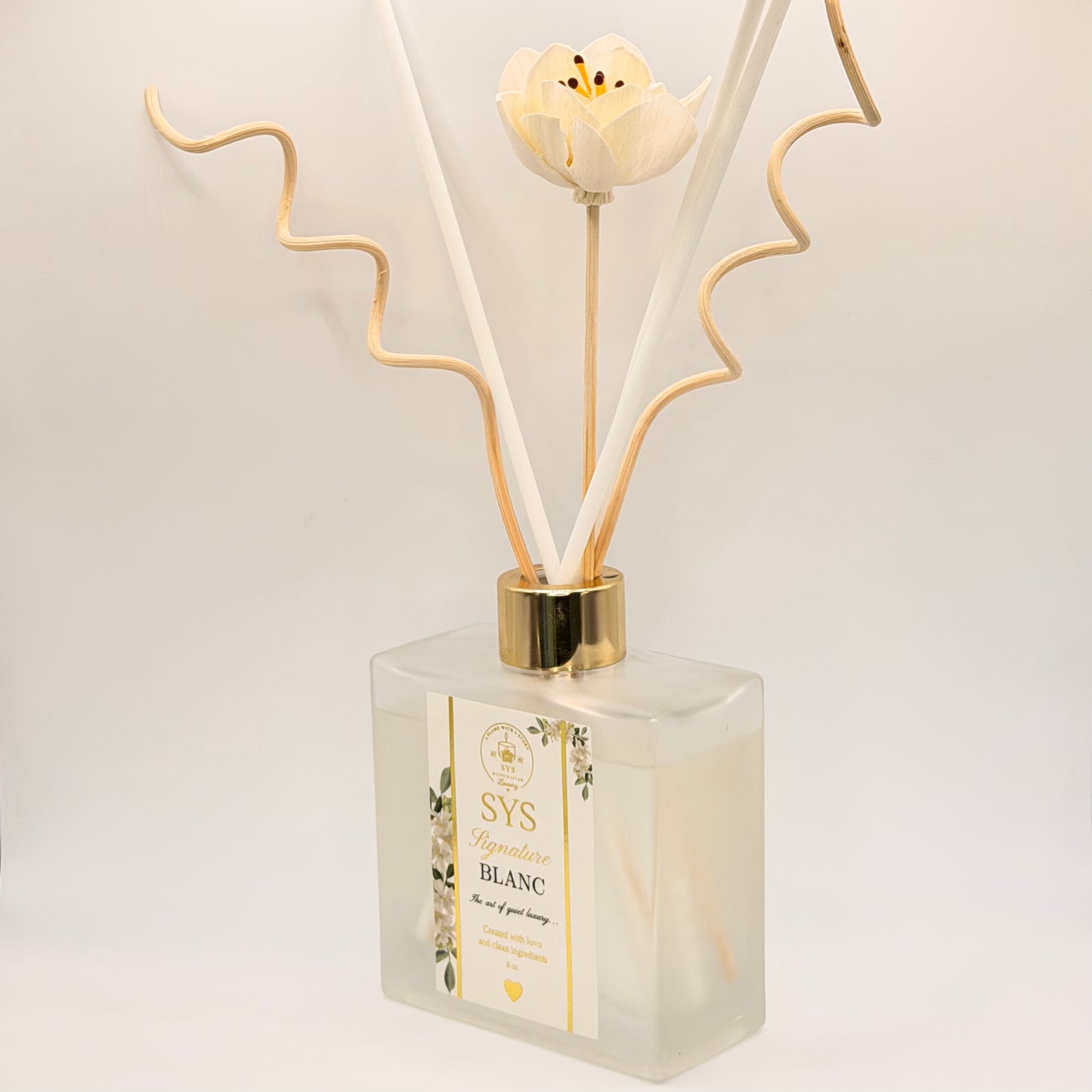 A reed diffuser bottle with a frosted glass container, a white flower cap, and beige reeds, shown on a clean white background. The label reads 'SYS Bougie Blanc'.