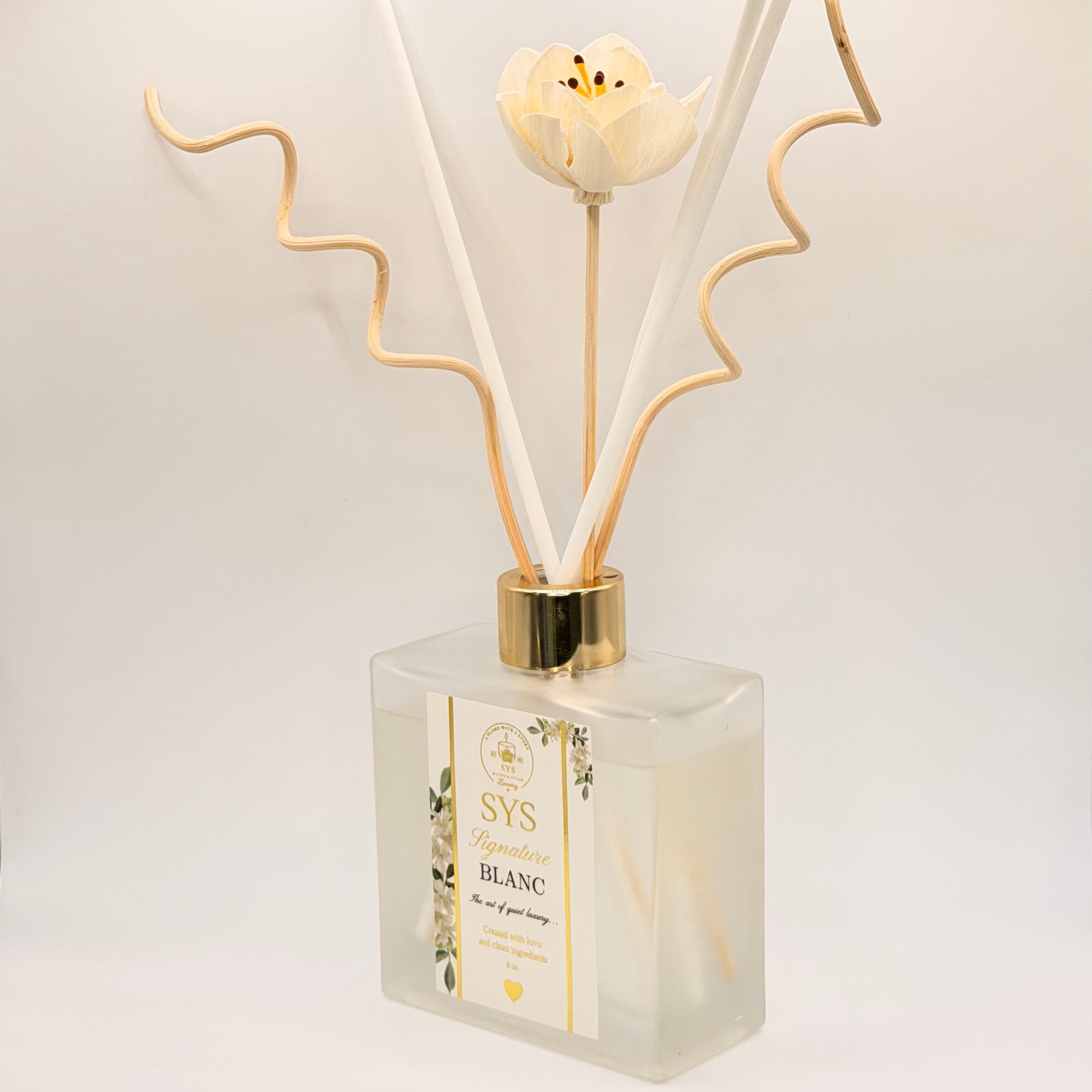 A reed diffuser bottle with a frosted glass container, a white flower cap, and beige reeds, shown on a clean white background. The label reads 'SYS Bougie Blanc'.