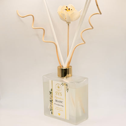 A reed diffuser bottle with a frosted glass container, a white flower cap, and beige reeds, shown on a clean white background. The label reads 'SYS Bougie Blanc'.