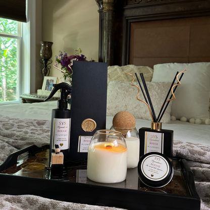 A styled bedroom scene with a black tray holding a lit white candle, a dark spray bottle, a round tin, and a reed diffuser, set on a bed in front of an ornate wooden headboard.