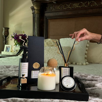 A styled bedroom scene showing a hand holding a reed diffuser stick over a tray with a candle, boxed diffuser, and other home fragrance items. The setting includes a dark wooden headboard, pillows, and a textured blanket.